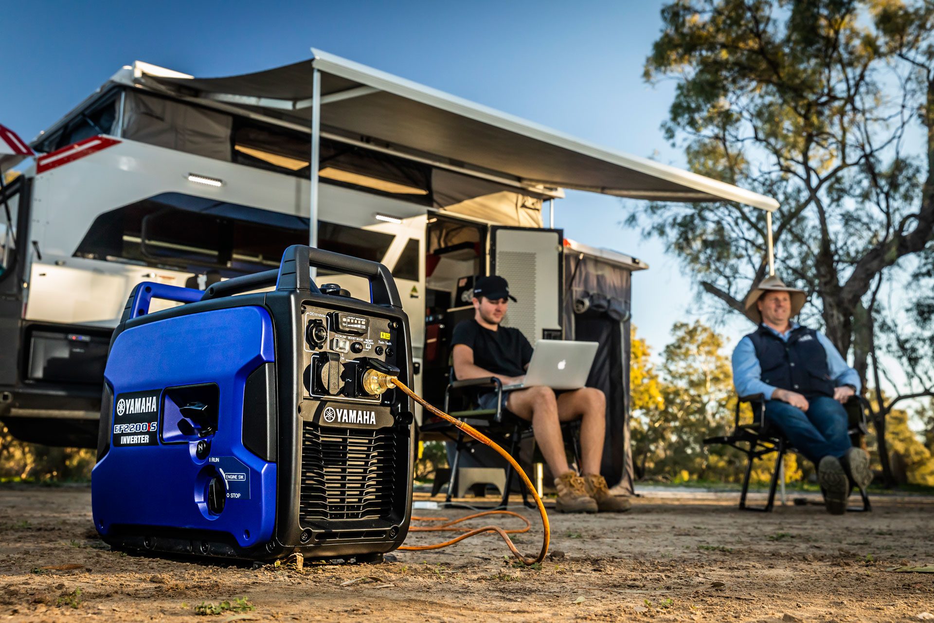 People working on a laptop plugged to a generator while sitting in front of camping trailer